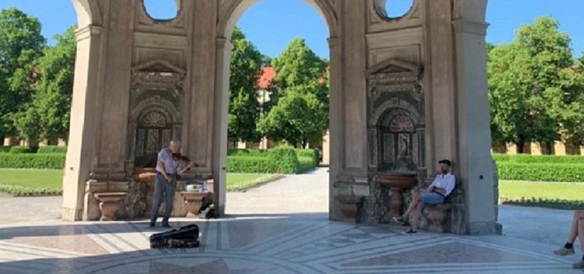 A man plays the violin in a park while another man sits on a bench and listens.